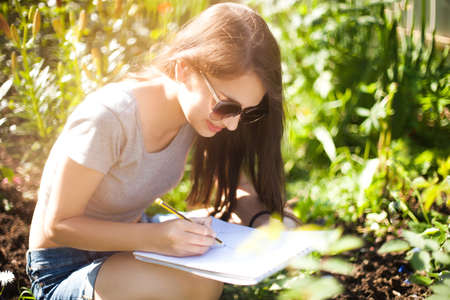 Attractive young artist works on plein air, drow in pencil. Wearing sunglasses, smiling. Enjoying painting on sunner day. Art education, art college.の写真素材