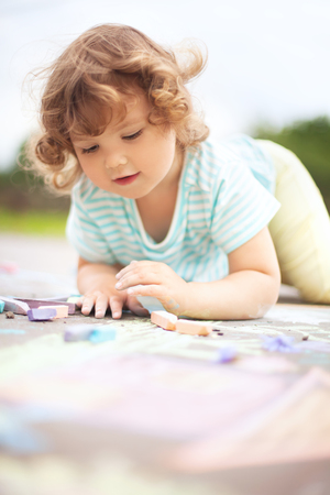 Sunny and warm summer days and happy chilhood. Cute little girl drawing with chalk crayons outdoorsの写真素材