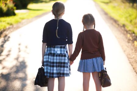 Adorable old-fashioned little 6 years old and 10 years old girls holding school bags. Preschooler and schoolgirl sisters, schoolkids are happy back to school. On the way to school, back view.の写真素材
