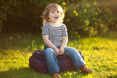 Adorable happy excited toddler girl with suitcase, waiting in the backyard. Tourism and adventure concept. Relocation with childrenの写真素材
