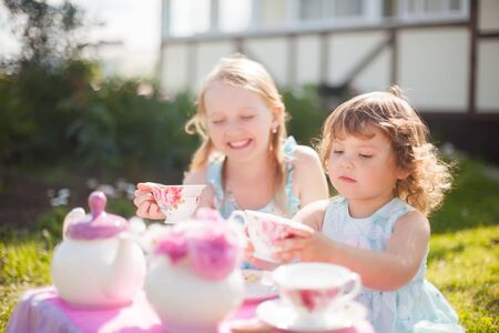 Adorable little sisters dressed like princesses playing tea party. Beautiful decorated tea party outdoors, at the backyard.の写真素材