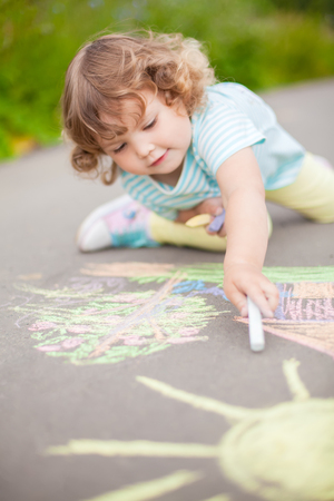Sunny and warm summer days and happy chilhood. Cute little girl drawing with chalk crayons outdoorsの写真素材