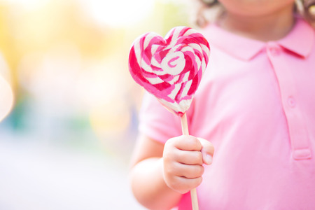 Beautiful little girl holding a big heart shaped lollipop, walking in the park, summer dayの写真素材