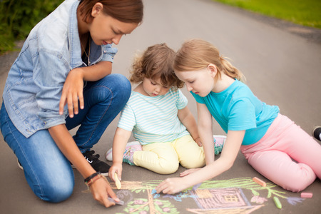 Little girls chalking at the asphalt with elder sister or nanny. Sisters friendship. Best friends children, happy family. Drawing with color chalk. Happy childhood. Kids in kindergarten. Preschooler leisure time. Kids spending time without parents. Children's drawings. Babysitting with two kids, young babysitter.の写真素材