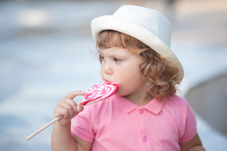 Beautiful little girl holding a big heart shaped lollipop, walking in the park, summer dayの写真素材