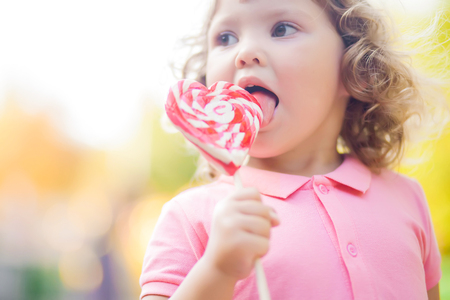 Beautiful little girl holding a big heart shaped lollipop, walking in the park, summer day.の写真素材