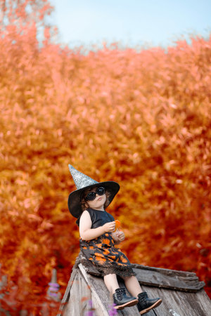 Little stylish witch outdoors, wearing halloween costume, ready for trick-or-treat, sitting on a...