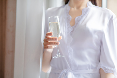 Beautiful young woman in white wedding robe holding champagne glass in hand. Bride drinking vine in the morning at the hotelの写真素材