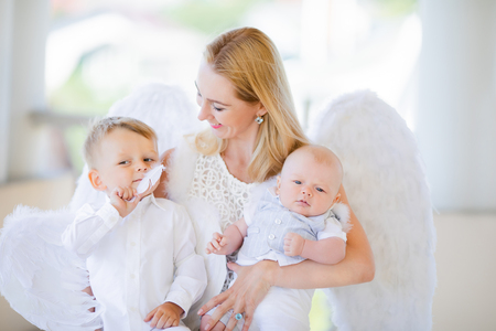 Beautiful mother with her toddler and newborn sons wearing angel costumes. Cheerful moment, loving family. Mom is the Guardian angel for her child concept.の写真素材