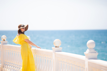 Lifestyle fashion portrait of happy young women, smiling and posing, wearing stylish sunglasses, straw hat and yellow dress, sea at the background.の写真素材