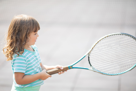 Child learning to play tennis on outdoor court. Little girl with tennis racket. Active exercise for kids.の写真素材