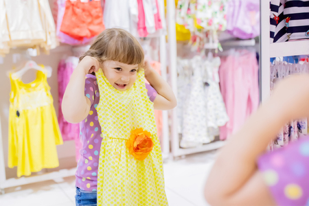 Adorable smiling little girl at the clothes store, try on new yellow summer dress, looking at the mirror.の写真素材