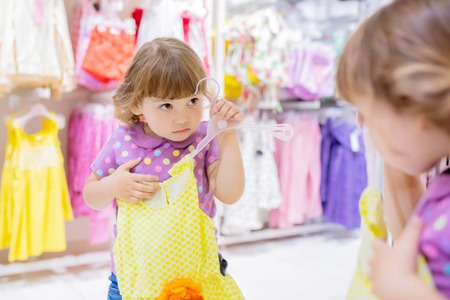 Adorable smiling little girl at the clothes store, try on new yellow summer dress, looking at the mirror.の写真素材
