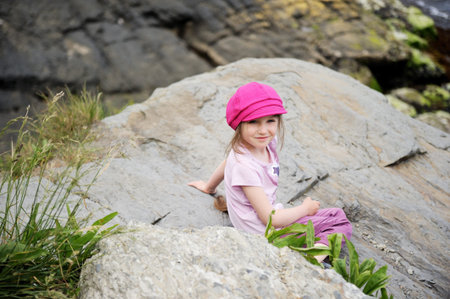 Happy little girl in pink sun hat sitting on the rocksの写真素材