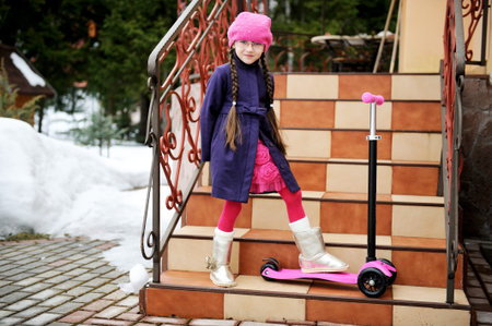 Toddler girl with pigtail in pink barret and purple coat posing with scooter on house stepsの写真素材