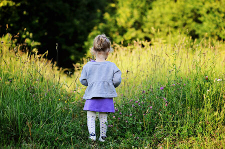 Adorable toddler girl with curly blond hair in knit purple dress and grey jacket playing outdoors on sunny dayの写真素材