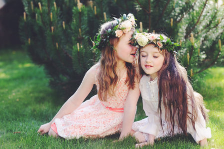 Two kid girls in elegant dresses with floral wreath on heads sitting on the lawn and speakingの写真素材