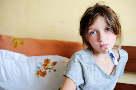 Ill child girl sitting in the bed and holding thermometrの写真素材