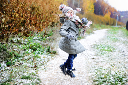 Adorable kid girl in grey coat hat and scarf walking in the fall forest with first snow