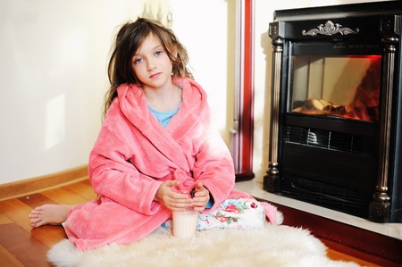 Adorable kid girl in pijama sitting near fireplace with glass of milkの写真素材