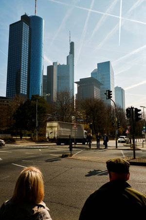 Frankfurt am Main, Germany - March 16, 2017: Elderly man and woman before the crossroad with truck and businessmen crossing the street with skyscrapers skyline on the background against the blue sky with planes contrailsのeditorial素材