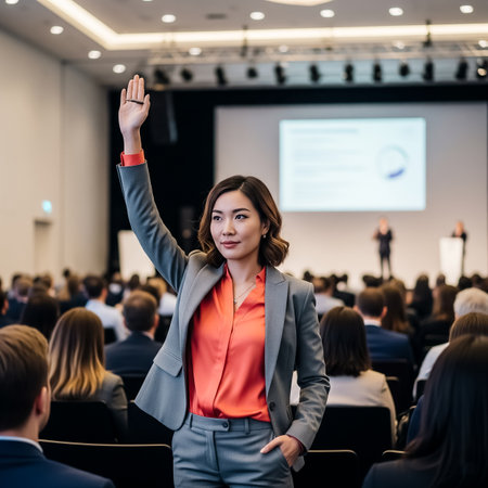 A professional woman in a gray suit and bright orange shirt raises her hand to ask a question during a conference. Attendees sit attentively, listening to speakers at the front.の素材