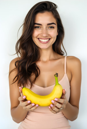 A cheerful woman stands in a well-lit indoor area, holding a ripe banana in both hands. Her smile radiates positivity, and the neutral background enhances the vibrant fruit.の素材