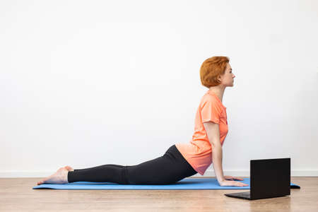 Yoga at home online. Quarantine. Coronavirus pandemic. Red-haired girl doing yoga asanas, looking at the laptop screen. Blue yoga mat. Against the background of a white wall.の写真素材