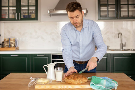 A man in a official shirt cuts freshly baked homemade bread on an oak table against a background of green kitchen. Quarantine, self-isolation. Antistress during the coronavirus pandemic. Copy space.の写真素材