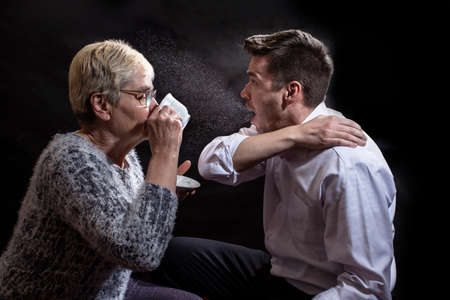 man coughs. Influenza, cold, coronavirus. Infection through an airborne droplet. The guy in the white shirt is coughing. Nearby, an elderly woman is drinking coffee from a cup.の写真素材