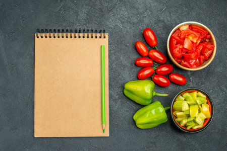 top view of bowls of bell pepper and tomatoes with vegetables and notepad on side on dark green...