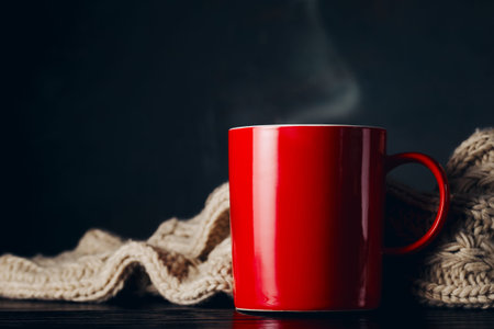 Composition of red mug with steam standing on table with warm knitted scarf lying near.