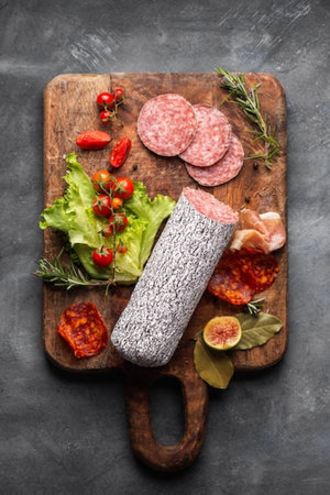 Sliced salami sausage on wooden cutting board with herbs and vegetablesの写真素材