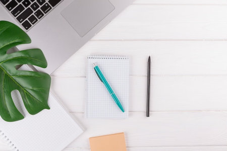 Top view of modern workspace with laptop, notepad and palm leaf on white wooden background