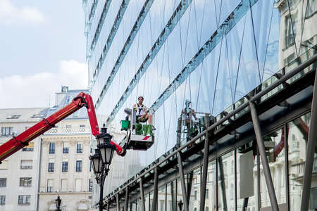 Budapest, Hungary - Aug 3, 2018: worker in elevated work platform cleaning building facade window glass by using equipment tools to cleanup dirty surface. Selective focusのeditorial素材