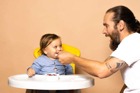 Dad laughs and feeds the baby on on the highchair. The concept of spending time with children.Front view photo on a light background. Life styleの写真素材