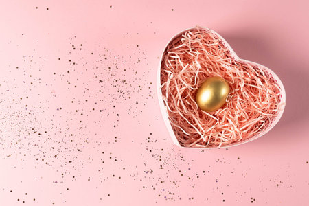Golden egg in a heart-shaped gift box on a pink background with gold sequins. A Easter egg in gift...