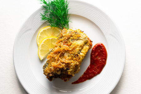 Battered fish on a white ceramic plate with lemon, red sauce and herbs on an isolated white...