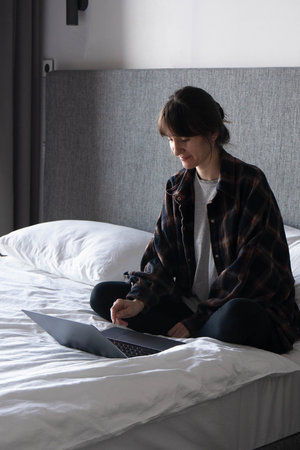 A young woman sits on the bed with laptop at home and pleasantly communicates with friends and...