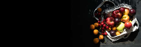 Fresh ripe fruits and berries on a black wooden background in an ecological mesh bag. Lifestyle. Hard shadows.Sunlight.Ripe harvest of apples. cherries, apricots and plums. Copy space.の写真素材