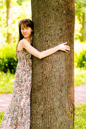 A young pretty girl in a long dress dreamily embraces a tree with her eyes closed.Ecology.Dreams. Financial growth. Reliability. Success. Investmentの写真素材