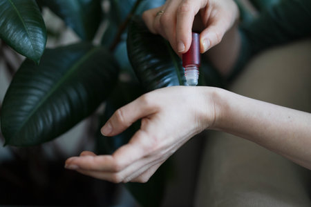 A gentle hand rolls essential oil onto the wrist in a serene indoor environment. Lush green leaves provide a calming backdrop, enhancing the sense of relaxation and wellness.の写真素材