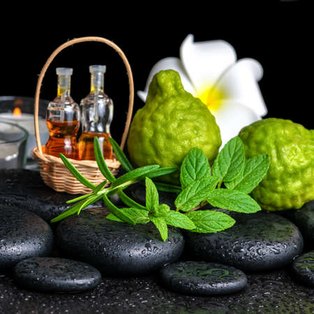 Aromatic spa of bottles essential oil in basket, fresh mint, rosemary, bergamot fruits, flower and candles on black zen stonesの写真素材