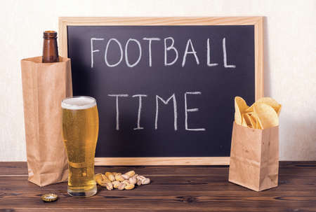 football fans setting of beer bottle in brown paper bag,  glass, chips, pistachio and handwriting text football time written in chalkboard over wooden tableの写真素材