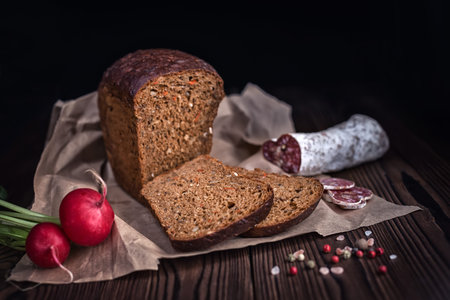 Siced brown bread, salami, radish and dry pepper on paper over wooden background