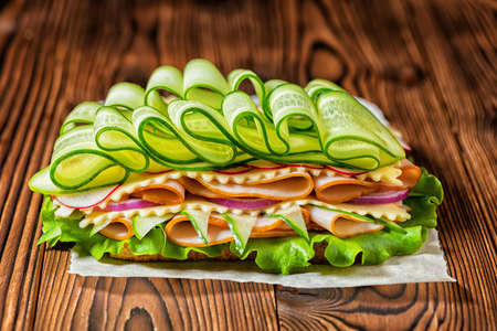 Rich sandwich with ham, cheese, bacon, radish, lettuce, cucumbers and onions on dark wooden background, concept of healthy eatingの写真素材