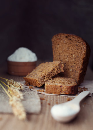 sliced brown bread with ears of wheat, flour or salt over dark wooden background, rustic style,...
