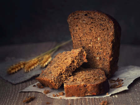 sliced brown bread with ears of wheat on paper over dark wooden background, rustic style, close up