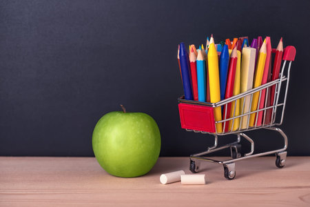 Education concept with green apple, chrome shopping cart, pens, crayons, chalks on black chalkboard...