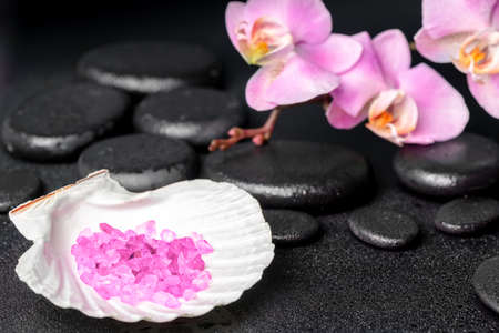 Beautiful spa setting of zen stones with drops, lilac orchid (phalaenopsis), cockshell with sea salt on black background, close upの写真素材